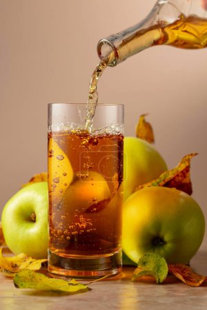 Photo for Apple juice is poured from a bottle into a glass. On a ceramic table ripe apples and dried-up leaves. - Royalty Free Image