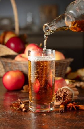 Foto de La sidra de manzana se vierte de una botella en un vaso. Bebida fresca con manzanas, canela y anís en una mesa de cocina. - Imagen libre de derechos