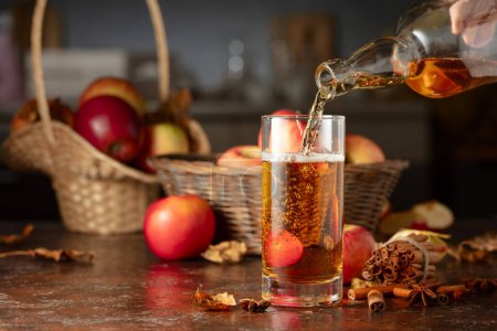 Photo for Apple cider is poured from a bottle into a glass. Fresh drink with apples, cinnamon, and anise on a kitchen table. - Royalty Free Image