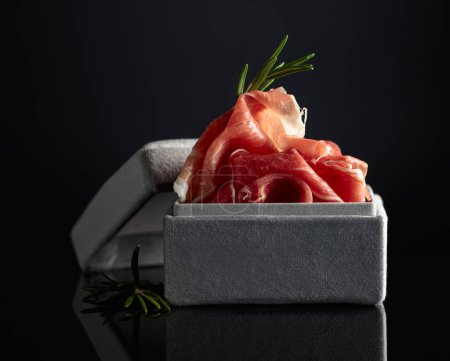 Téléchargez les photos : Prosciutto au romarin dans une boîte cadeau grise sur fond noir. Concept du thème de la nourriture chère. - en image libre de droit