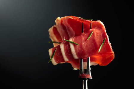 Photo for Sliced prosciutto with rosemary on a fork, black background, copy space. - Royalty Free Image