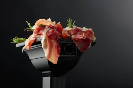 Photo for Prosciutto with rosemary on a black podium. Copy space. - Royalty Free Image