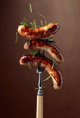 Photo for Grilled Bavarian sausages with rosemary.  Sausages  on a fork sprinkled with rosemary. Brown background. - Royalty Free Image