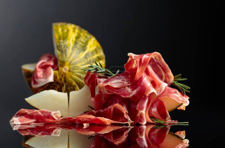 Photo for Prosciutto with melon and rosemary on a black reflective background. - Royalty Free Image
