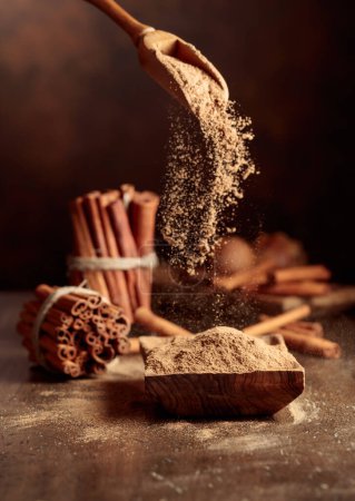 Photo for Cinnamon powder is poured into a wooden bowl. In the background are kitchen utensils and cinnamon sticks. - Royalty Free Image