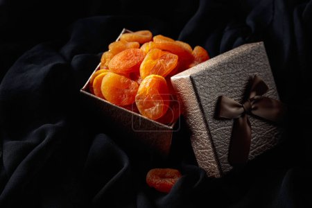 Téléchargez les photos : Abricots secs dans une boîte cadeau sur un chiffon noir. - en image libre de droit