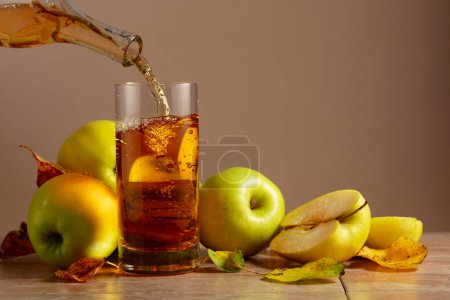 Téléchargez les photos : Le jus de pomme est versé d'une bouteille dans un verre. Sur une table en céramique des pommes mûres et des feuilles séchées. Fond beige avec espace de copie. - en image libre de droit