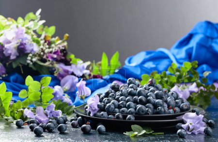 Photo for Fresh juicy blueberries on a black plate. Summer still life with blueberries, colored sweet peas and meadow grasses on a dark blue background. - Royalty Free Image