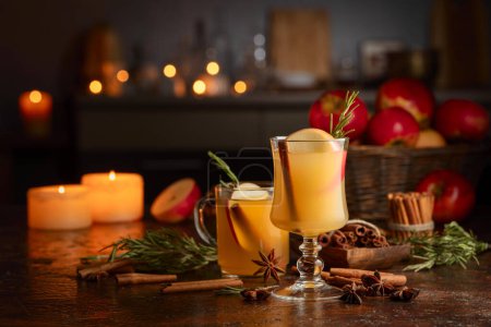 Photo for Mulled cider with slice apples, cinnamon, rosemary, and anise stars in glass cups on a brown rustic table. Holiday traditional hot drink. On a dark background kitchen utensils and burning candles. - Royalty Free Image