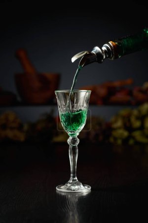 Foto de Licor de hierbas verdes se vierte de una botella vintage en un vaso. En el fondo hierbas secas y utensilios de cocina viejos. - Imagen libre de derechos
