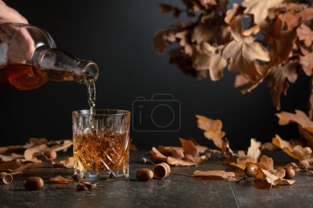 Téléchargez les photos : Le whisky est versé d'une bouteille dans un verre. Whisky sur une table noire avec des feuilles de chêne séchées. - en image libre de droit