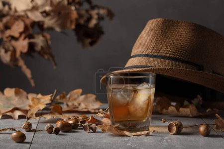 Foto de Whisky con hielo sobre una mesa de piedra con hojas secas de roble y sombrero de hombre. - Imagen libre de derechos