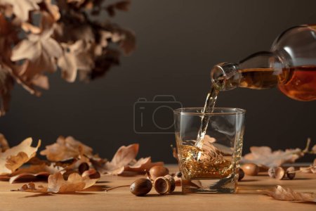 Téléchargez les photos : Le whisky est versé d'une bouteille dans un verre. Whisky sur une table en bois avec des feuilles de chêne séchées. - en image libre de droit
