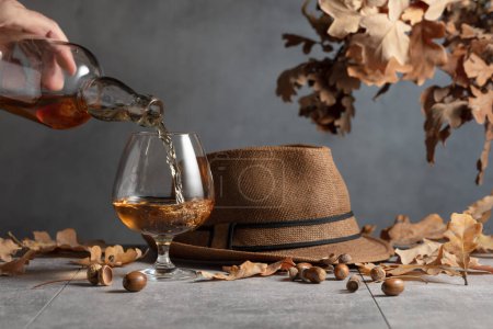 Téléchargez les photos : Le brandy est versé d'une bouteille dans un verre. Snifter de cognac sur une table en pierre aux feuilles de chêne séchées. - en image libre de droit