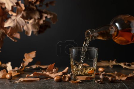 Foto de El whisky se vierte de una botella en un vaso. Whisky sobre una mesa negra con hojas secas de roble. - Imagen libre de derechos