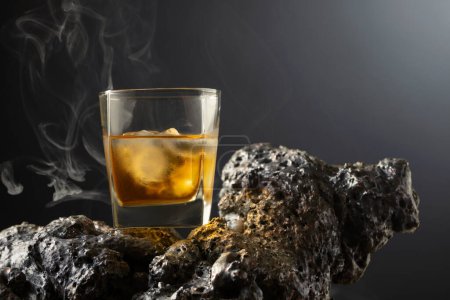 Foto de Whisky con hielo y humo en una piedra gris. Fondo negro con espacio de copia. - Imagen libre de derechos