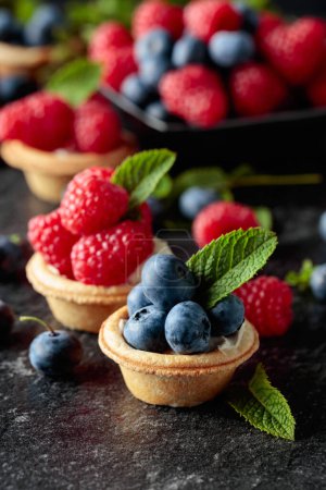 Photo for Small tartlets with fresh raspberries and blueberries garnished with mint on a black background. - Royalty Free Image
