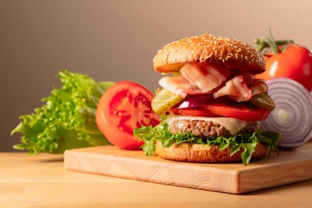 Téléchargez les photos : Hamburger frais savoureux sur une planche à découper en bois. Burger à la tomate, à l'oignon, au concombre en conserve, à la salade, au fromage, à l'escalope de bœuf et au bacon. Espace de copie. - en image libre de droit