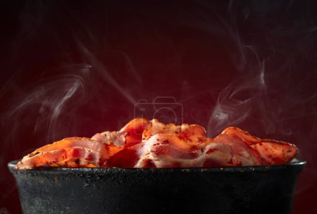Photo for Fried steaming bacon slices in an old black pan on a dark red background. Copy space. - Royalty Free Image