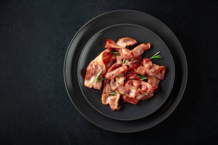 Photo for Roasted bacon slices with rosemary on a black plate. Copy space. Top view. - Royalty Free Image