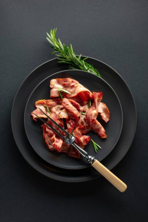 Téléchargez les photos : Tranches de bacon rôties avec romarin sur une assiette noire. Espace de copie. Vue du dessus. - en image libre de droit