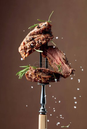Photo for Grilled beef steak with rosemary on a brown background. Beef steak on a fork sprinkled with rosemary and sea salt. - Royalty Free Image