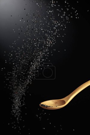 Photo for Grains of black sesame is poured with a wooden spoon. Black sesame on a dark background. Copy space. - Royalty Free Image