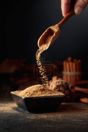 Photo for Cinnamon powder is poured into a black bowl. In the background are kitchen utensils and cinnamon sticks. - Royalty Free Image