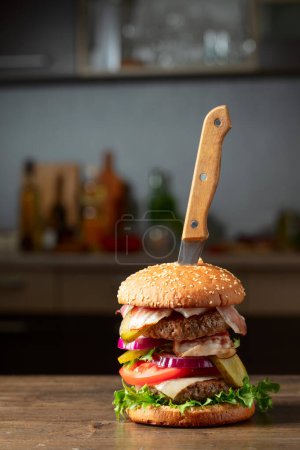 Photo for Fresh burger with knife on a kitchen table. Burger with tomato, onion, preserved cucumber, salad, cheese, beef cutlet, and bacon. - Royalty Free Image