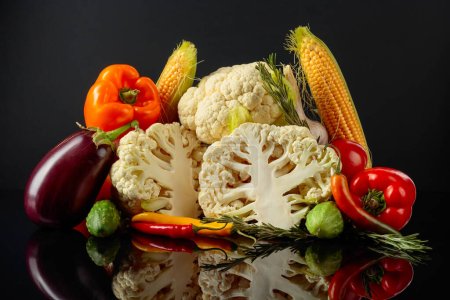 Foto de Varias verduras frescas sobre un fondo negro reflectante. Se presentan tomates, pimentón, coliflor, ají, ajo, berenjena, pepinos, maíz y romero.. - Imagen libre de derechos