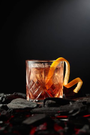 Foto de Cóctel a la antigua con hielo y piel de naranja. Un vaso con un cóctel sobre carbón quemado. Copiar espacio. - Imagen libre de derechos