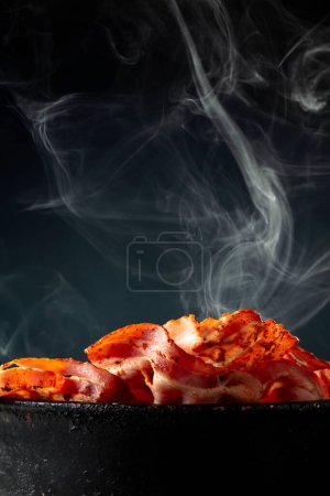Téléchargez les photos : Tranches de bacon cuites à la vapeur dans une vieille casserole noire sur fond noir. Espace de copie. - en image libre de droit