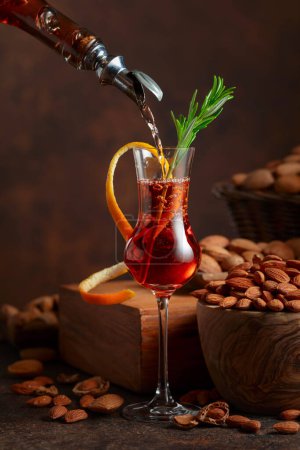 Foto de Licor italiano Amaretto se vierte de una botella en un vaso. Vaso de licor decorado con ramita de romero y piel de naranja. - Imagen libre de derechos