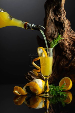 Téléchargez les photos : Liqueur italienne traditionnelle Limoncello sur fond noir réfléchissant. Verre de liqueur de citron avec brindille de romarin sur un fond de vieux chicots. L'alcool est versé dans un verre. - en image libre de droit