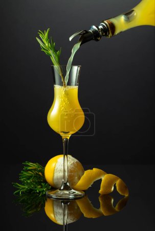 Foto de Licor tradicional italiano Limoncello con ramita de romero sobre fondo negro reflectante. Licor se vierte de una botella en un vaso. - Imagen libre de derechos