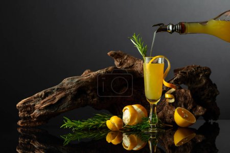 Photo for Traditional Italian liqueur Limoncello on a black reflective background. Glass of lemon liquor with rosemary twig on a background of old snag. Liquor is poured into a glass. - Royalty Free Image
