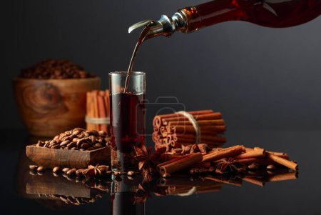 Photo for Coffee liquor is poured from a bottle into a glass. Coffee beans, cinnamon sticks, and anise are scattered on the black reflective background. - Royalty Free Image