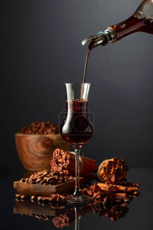 Photo for Coffee liquor is poured from a bottle into a glass. Coffee beans, cinnamon sticks, and anise are scattered on the black reflective background. - Royalty Free Image