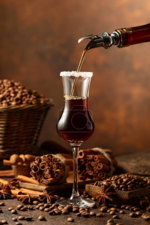 Téléchargez les photos : La liqueur de café est versée d'une bouteille dans un verre. Les grains de café, les bâtonnets de cannelle et l'anis sont éparpillés sur une table vintage. - en image libre de droit