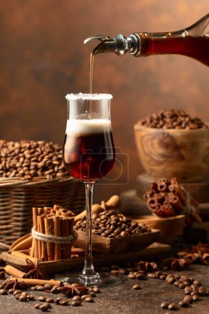 Foto de El licor de café se vierte de una botella en un vaso con crema. Los granos de café, los palitos de canela y el anís están esparcidos en la mesa. - Imagen libre de derechos