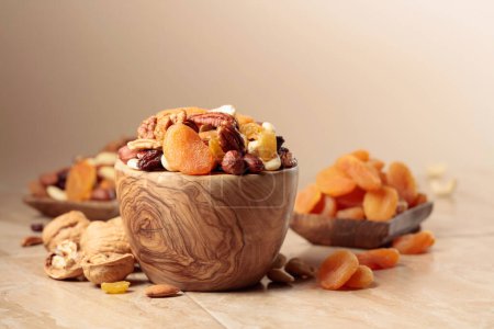 Téléchargez les photos : Dried fruits and nuts on a beige ceramic table. The mix of nuts, apricots, and raisins in a wooden bowl. Copy space. - en image libre de droit