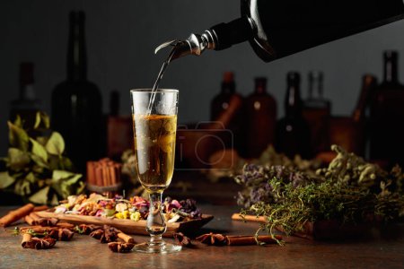Photo for Yellow herbal liquor or mixture is poured from a vintage bottle into a glass. On a table dried herbs, flowers, spices, and old kitchen utensils. Concept of herbal medicine. - Royalty Free Image