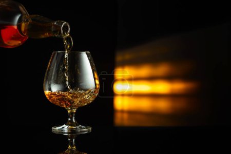 Photo for Pouring brandy from a bottle into a snifter on a black reflective background. Copy space. - Royalty Free Image