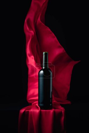 Foto de Botella de vino tinto y aleteos de tela roja sobre fondo negro. - Imagen libre de derechos