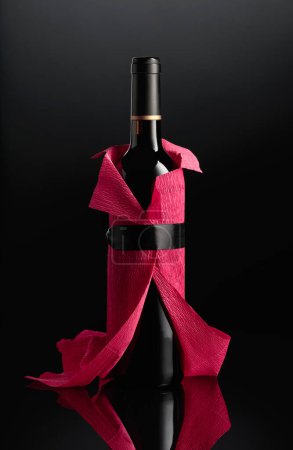Téléchargez les photos : Bottle of red wine wrapped in crepe paper on a black background. The bottle looks like a woman in a red evening dress. - en image libre de droit
