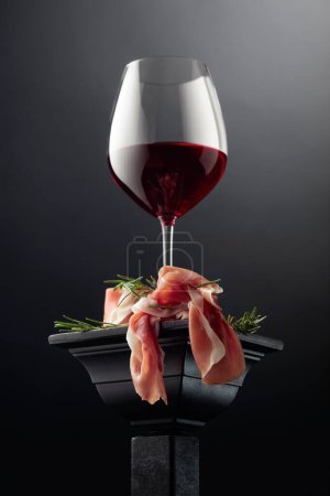 Photo for Glass of red wine and prosciutto with rosemary on a black podium. - Royalty Free Image