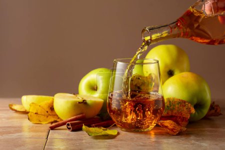 Téléchargez les photos : Le jus de pomme est versé d'une bouteille dans un verre. Sur une table en céramique, pommes mûres, bâtonnets de cannelle et feuilles séchées. Fond beige avec espace de copie. - en image libre de droit