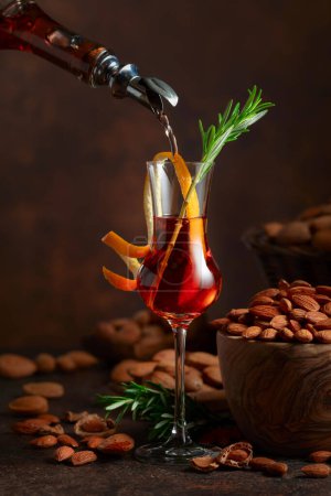 Photo for Italian liqueur Amaretto is poured from a bottle into a glass. Glass of liqueur garnished with rosemary twig and orange peel. - Royalty Free Image
