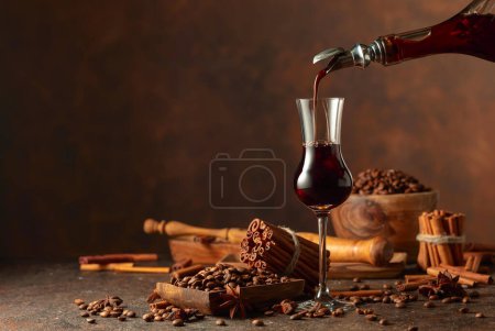 Photo for Coffee liquor is poured from a bottle into a glass. Coffee beans, cinnamon sticks, and anise are scattered on the vintage table. Copy space. - Royalty Free Image