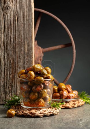 Foto de Aceitunas picantes en un tazón de vidrio. Cuenco con aceitunas conservadas y ramitas de romero sobre una mesa de piedra. - Imagen libre de derechos